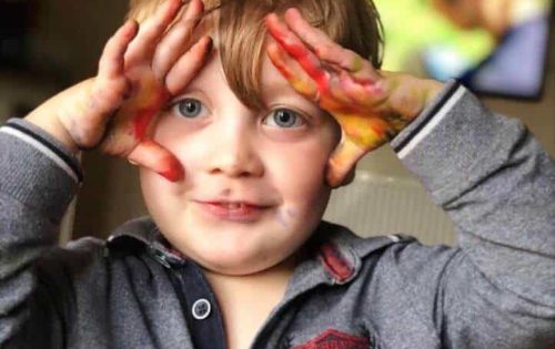 The image is of a young boy holding his hands up towards his head, with paint on his hands doing messy fun play for sensory processing challenges.