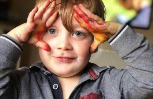 The image is of a young boy holding his hands up towards his head, with paint on his hands doing messy fun play for sensory processing challenges.