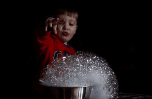 The image is of a dark bowl full of bubbles, with a child stood behind holding some of the bubbles up in the air.