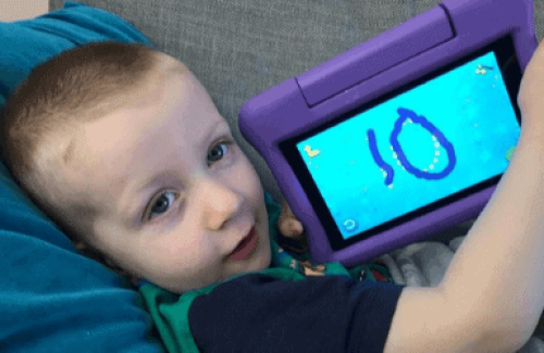 An image of a young boy looking into the camera holding up a console device screen, with the number ten written on the screen.