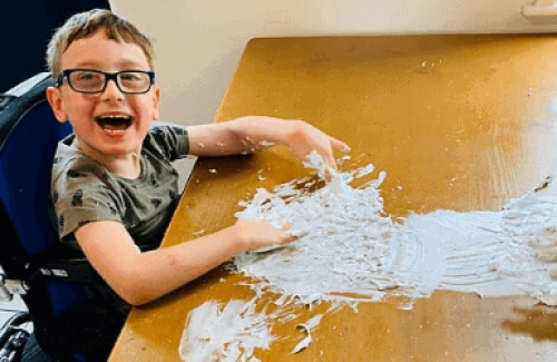 The image is of a young wheelchair user, sat up at a table, engaged in messy play with some dough out in front of him on the table.