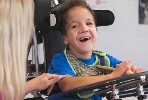 The circular image is of a young child from the waist up smiling at an adult in a piece of equipment that is assisting him to stand.