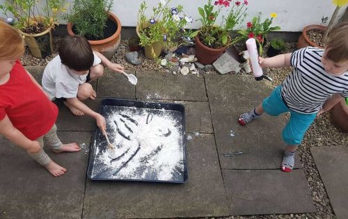 The image is of three children out on a patio, in a garden, two stood and one bending down engaging in a messy tray for play with lots of flour.