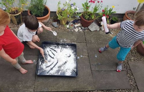 The image is of three children out on a patio, in a garden, two stood and one bending down engaging in a messy tray for play with lots of flour.