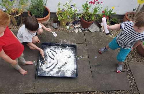 The image is of three children out on a patio, in a garden, two stood and one bending down engaging in a messy tray for play with lots of flour.