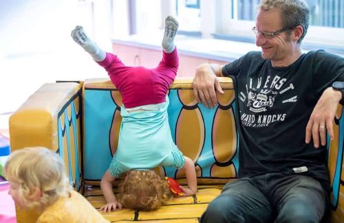 An image showing a man sat down next to two children, one on the ground, the other doing a headstand inside a soft play pirate ship.