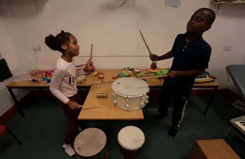The image is of two children stood around a table with various instruments, including drums, mid dance, about to bang the drums with their drumsticks looking at each other.