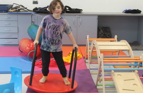 A child with a big smile stands confidently on a red balance board with handrails, wearing a colorful tie-dye shirt that reads 'Give Me Space.' The room is set up with vibrant mats, sensory equipment, and wooden climbing frames, creating an engaging and inclusive play environment