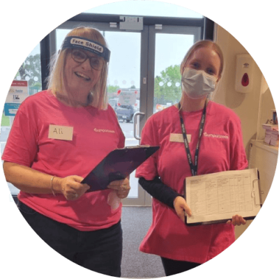 Two women in obright tops stood at Gympanzees Pop Up reception, holding clipboards with a masks on.
