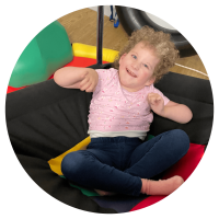 A young girl sat cross-legged in a seated small hammock-like sling smiling at the camera.