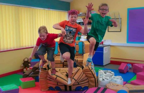 The image is of 3 young boys, all jumping off the edge of a soft play pirate ship, surrounded by soft play apparatus.